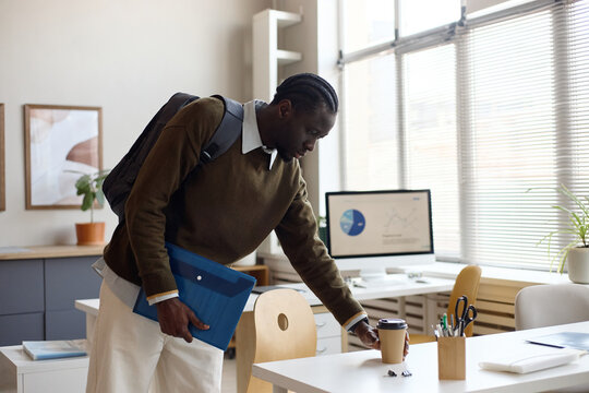 Black young adult man arriving at office, setting takeaway coffee on desk while carrying backpack and document folder. Ideal for workplace routine, onboarding, commuting, productivity themes
