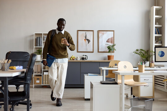 Wide shot of Black young adult man walking through office, carrying folder and takeaway coffee, arriving for work. Useful for workplace routine, employee onboarding, corporate lifestyle marketing