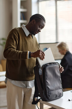 Vertical shot of Black young adult man packing laptop into backpack in office, preparing for workday or commute. Suitable for remote work, hybrid work, business mobility marketing