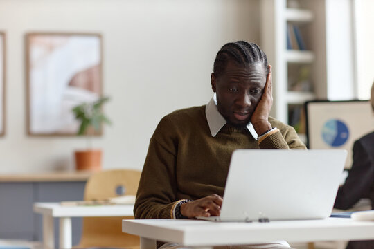 Black young adult man working on laptop while holding face, showing stress or fatigue during office task. Useful for burnout, workload pressure, productivity, mental health themes