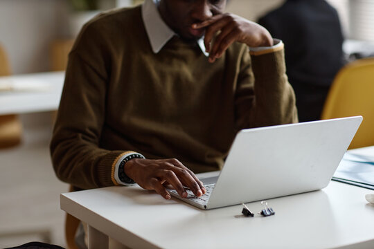 Medium close up shot of Black young adult man working on laptop, thinking through task in office setting. Suitable for business productivity, remote work, planning, or corporate training use