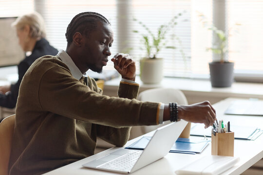 Young Black businessman working at laptop, reaching for pen while planning tasks at office desk. Suitable for corporate productivity, workflow, planning, or office routine marketing
