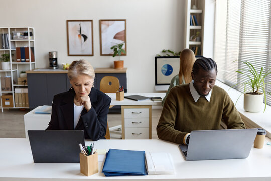 Senior Caucasian businesswoman and young Black businessman working on laptops in shared office, supporting teamwork and productivity. Useful for corporate communication, hybrid work