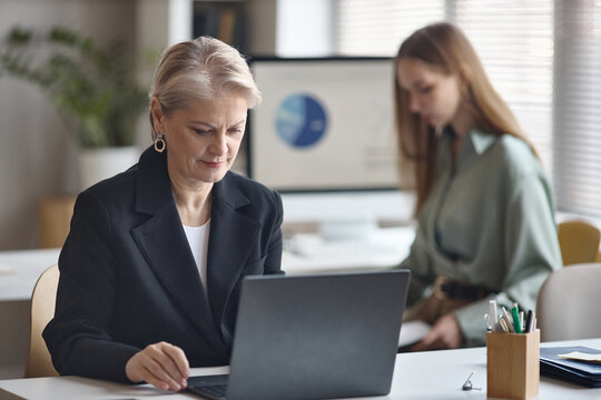 Senior businesswoman working on laptop in office while young female colleague reviewing data in background. Suitable for corporate teamwork, leadership, finance, strategy marketing