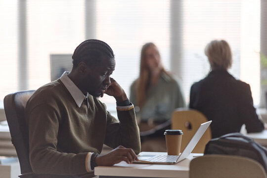 Medium shot of Black young adult man working on laptop in office, resting head on hand and focusing on task. Suitable for remote work, productivity, burnout, or corporate planning campaigns