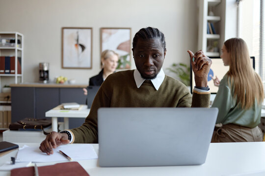 Portrait of Black young adult man working on laptop in office, raising hand while discussing task with coworkers. Suitable for teamwork, corporate communication, productivity campaigns