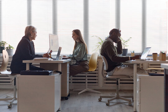 Wide shot of diverse office team working at desks, discussing tasks, using computers in workplace. Suitable for business communication, teamwork, productivity marketing