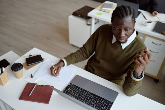 High angle shot of Black young adult businessman procrastinating at desk beside laptop, looking away. Useful for burnout, productivity, planning
