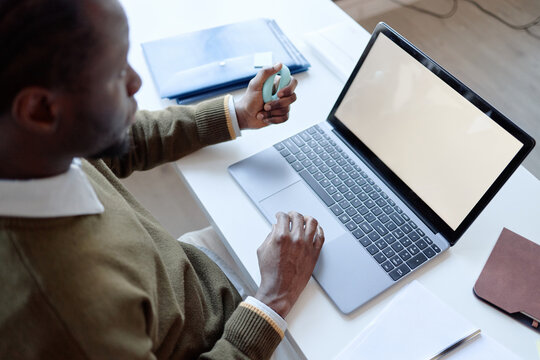 High angle view of Black young adult man using laptop while holding fidget toy at desk, managing workflow. Ideal for hybrid work, tech support, productivity marketing