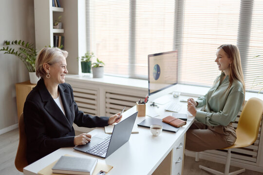 Senior businesswoman meeting young adult colleague in office, discussing project data on laptop and desktop monitor. Ideal for teamwork, mentoring, leadership, workplace communication