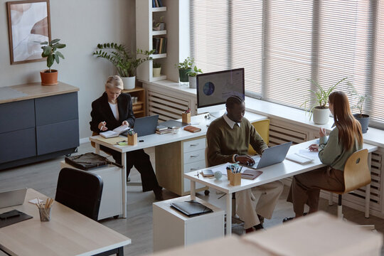 Wide angle shot of diverse office team working at desks, typing on laptops and reviewing notes during meeting. Supporting business collaboration, corporate workflow, hybrid work marketing