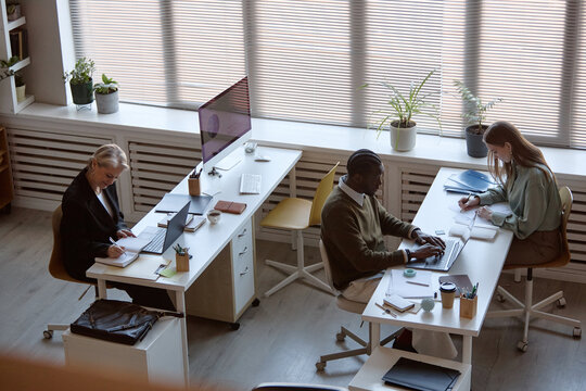 High angle shot of diverse office team working at desks, typing on laptop, writing notes, reviewing documents. Supporting business workflow, teamwork, productivity or corporate communication use