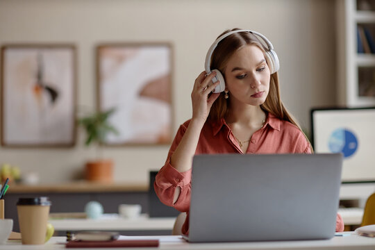 Young adult woman wearing headphones, working on laptop and adjusting audio during online meeting or training. Ideal for office work, e learning, productivity, digital communication