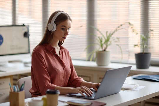 Young adult woman wearing headphones working on laptop at desk, managing online communication and focused on workflow. Ideal for office work, customer support, e learning