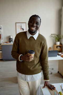 Vertical shot of Portrait of Black young adult man holding takeaway coffee and smiling at camera in office. Supporting workplace branding, employee wellbeing, recruitment, corporate culture marketing