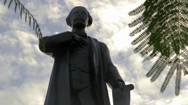 Statue of Juan Pablo Duarte, founding father of the Dominican Republic, seen from low angle against dramatic cloudy sky and tropical foliage.