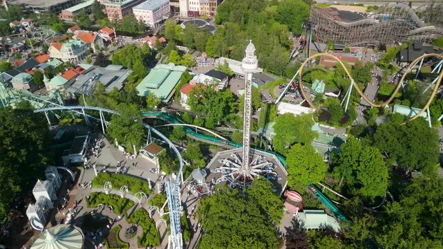 Drone orbits packed Liseberg park revealing tall drop tower, spinning rides, wooden roller coaster and ornate carousel amid lush Gothenburg summer cityscape