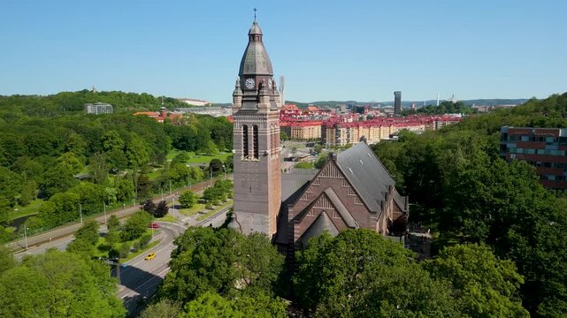 Aerial drone parallax shot of Annedalskyrkan's brick clock tower rising above lush summer trees with Gothenburg's red-roofed cityscape spreading in background