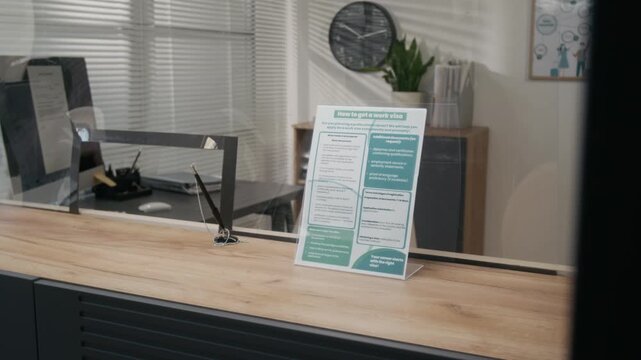Medium no people shot of applicant service counter at visa center, embassy or consulate, with glass partition, computer on officer desk, information poster with work permit instructions