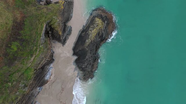 Aerial view of the rugged coastline where dark rocks meet the sandy beach and turquoise sea, creating a stunning contrast of textures and tones, Fowey, Cornwall, United Kingdom.