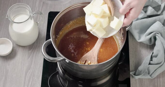 Closeup of adding butter in pot with sugar on stove  stirring with wooden spoon to make homemade salted caramel sauce on gray kitchen table. Cooking process caramel made from sugar, butter, cream