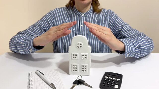 Female hands forming protective roof over house model at office desk with calculator keys home insurance concept