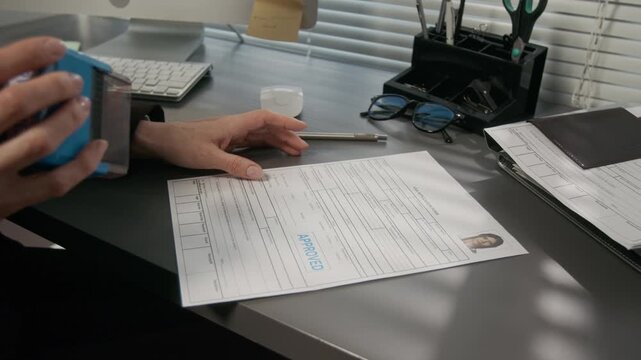 Medium close-up of hands of unrecognizable female embassy officer putting approval stamp on study visa application of young woman, stack of passports, stationery and computer on desk