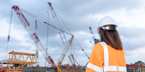 A worker stands on a construction site. Several cranes are in operation, lifting materials. The sky is cloudy, and the worker looks at the ongoing tasks with a focus on safety © Happy Photo