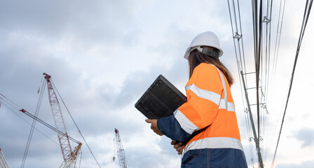 A construction worker stands on a site holding a tablet. Cranes are visible in the background among power lines. The sky is cloudy and the work continues © Happy Photo