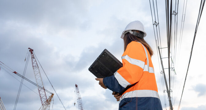 A construction worker stands on a site holding a tablet. Cranes are visible in the background among power lines. The sky is cloudy and the work continues