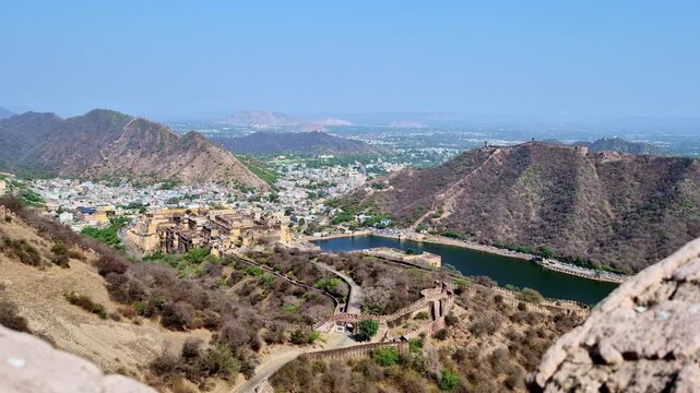 Showing a scenic view of Jaigarh Fort, Maota Lake, and the surrounding Aravalli hills captured between large rocky foreground stones from the top of Nahargarh Fort, with the city visible.