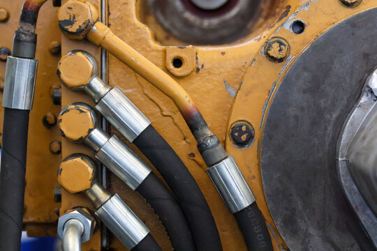 Close-up of industrial hydraulic system showing black high-pressure hoses with metal fittings and bolts connected to a yellow heavy machinery engine block outdoors.