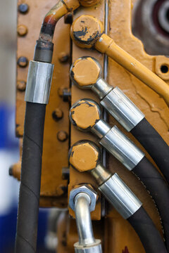 Close-up of industrial hydraulic system showing black high-pressure hoses with metal fittings and bolts connected to a yellow heavy machinery engine block outdoors.