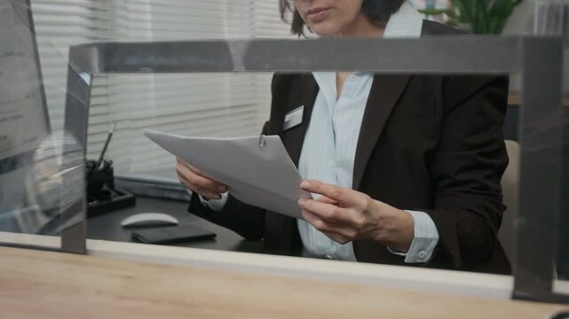 Medium zoom-out shot of middle-aged Caucasian female visa center agent checking documents submitted for visa application, while sitting at customer service counter behind glass partition