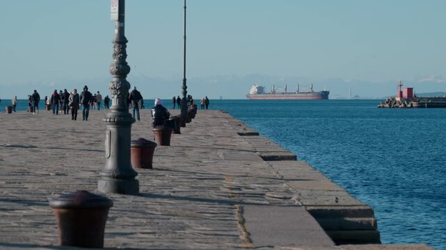 Wide view of people walking on Molo Audace pier in Trieste Italy with historic lamp post bollards and a large cargo ship on the horizon with snowy Alps backdrop