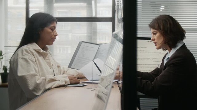 Side shot of middle-aged Caucasian female visa center employee in business suit consulting Hispanic client on paperwork, pointing to mistakes in application form during document verification