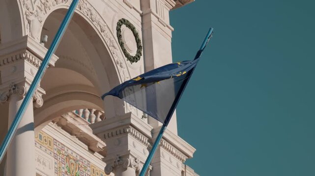 Close up of the European Union flag waving in the wind in front of the ornate architecture and arches of a historic building in Piazza Unita d Italia Trieste