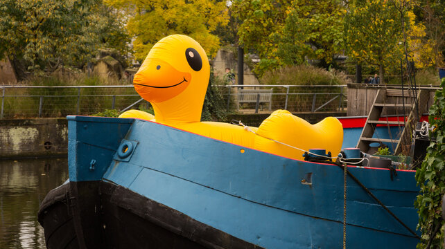 A large, inflatable yellow duck sitting on a blue wooden barge on a canal in Amsterdam, Holland