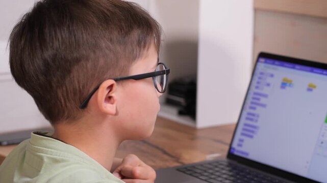 Young boy with glasses focused on a laptop screen, learning to program in an educational setting, developing his computer science skills 