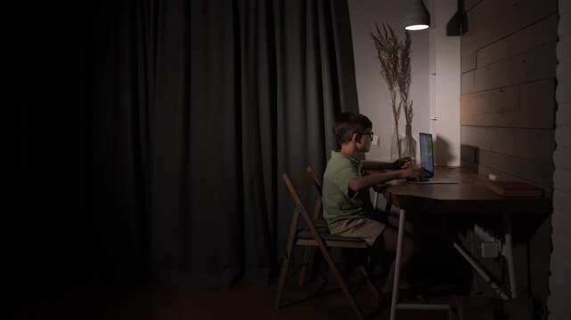 Young boy wearing glasses concentrating on his schoolwork or online learning, typing on a laptop computer at a wooden desk under a focused lamp light late in the evening