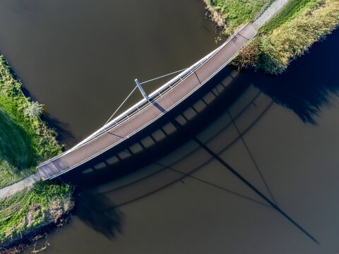 Aerial view of a pedestrian bridge casting long shadows on the dark water, contrasting with the green banks on either side, Almere, Flevoland, Netherlands.