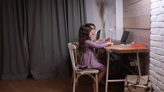 Boy and girl sitting at a table together, actively engaged in online learning with a laptop, exploring sustainability and alternative electric power sources using a small wind turbine model