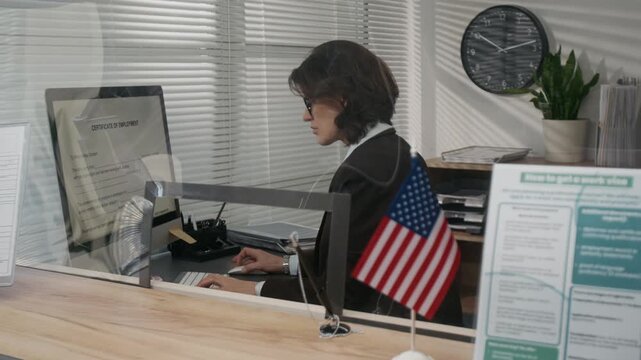 Medium side shot of busy female American consulate visa agent working with documents on computer behind glass partition, USA flag next to information leaflet on applying for work permit