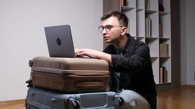 Man focusing on his laptop while typing, balancing it on stacked suitcases, representing remote work, digital nomad lifestyle, and business travel preparations