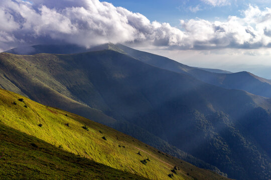 Mountain landscape rolling green hills dramatic clouds casting shadows sunlit slopes.