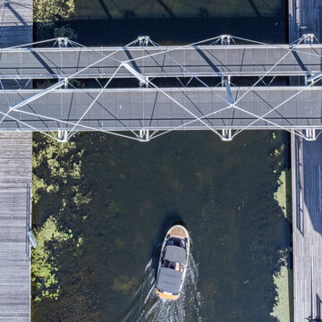Aerial view of a boat sailing under a modern bridge, its wake contrasting against the dark water and patches of algae, Hengelostraat, Almere, Flevoland, Netherlands.
