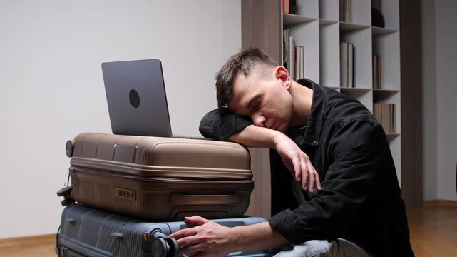 Young man feeling exhausted and sleeping on his stacked suitcases with a laptop as he experiences travel burnout, waiting for departure in an airport or transit lounge