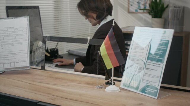 Medium shot of busy Caucasian female official in formal jacket processing work visa application at consulate, checking personal information and typing on computer, German flag and leaflet on counter