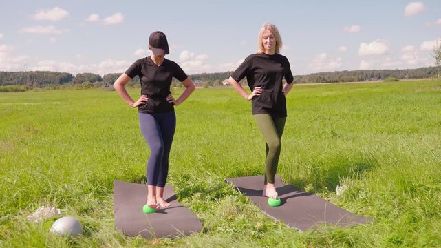Two active women performing foot massage and proprioception exercises with hemisphere balance trainers on yoga mats in a sunlit green field, improving coordination and strengthening ankle muscles