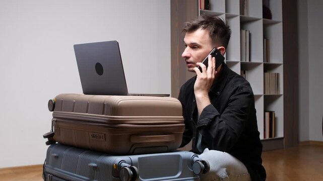 Man working on a laptop placed on stacked suitcases while talking on a mobile phone, balancing travel and professional duties, embodying the challenges of modern remote work and business travel